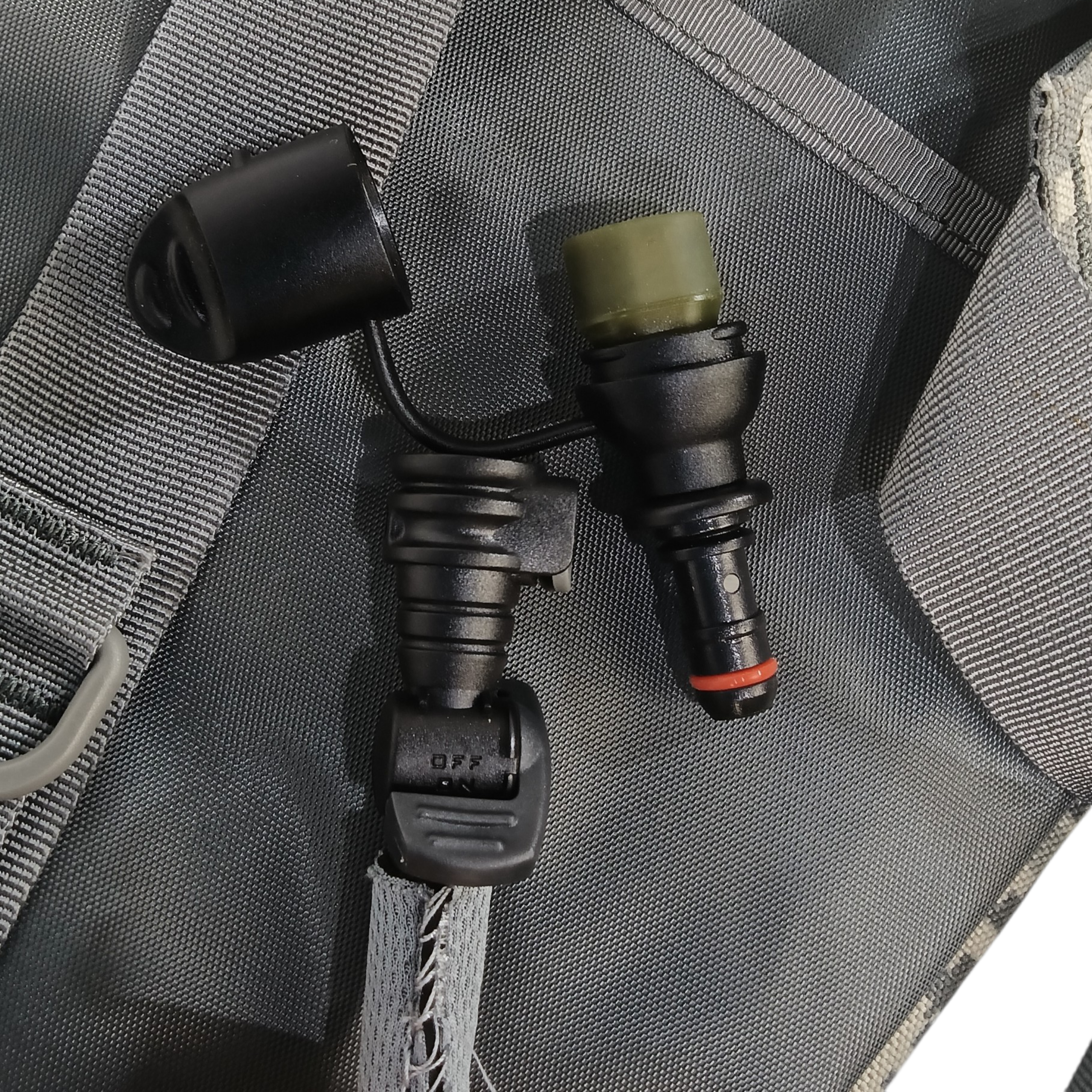 Sac d'hydratation 3 Litres ACU: bouchons et valves en noir et vert kaki près d'une fermeture éclair sur un sac gris foncé.
