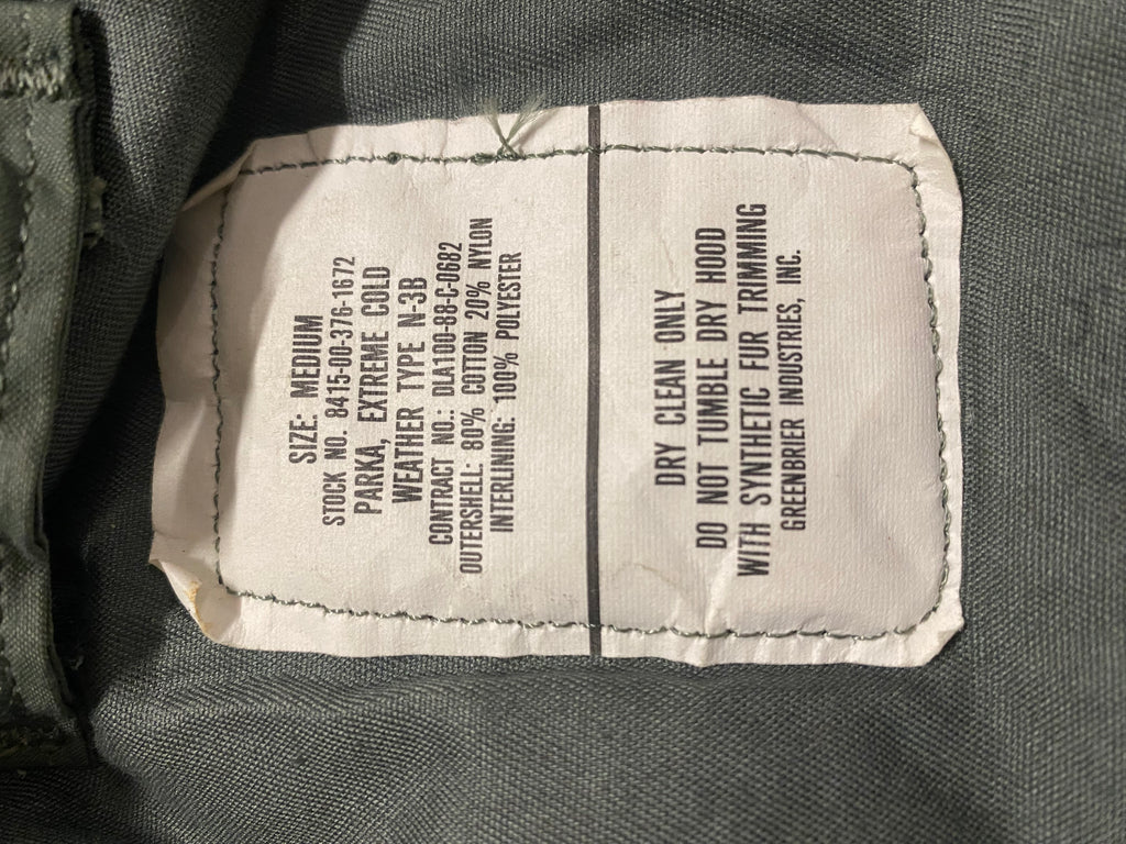 Parka N3B US Army grand froid extrême authentique: étiquette textile blanche cousue sur tissu olive gris, détails de composition et instructions d’entretien visibles.