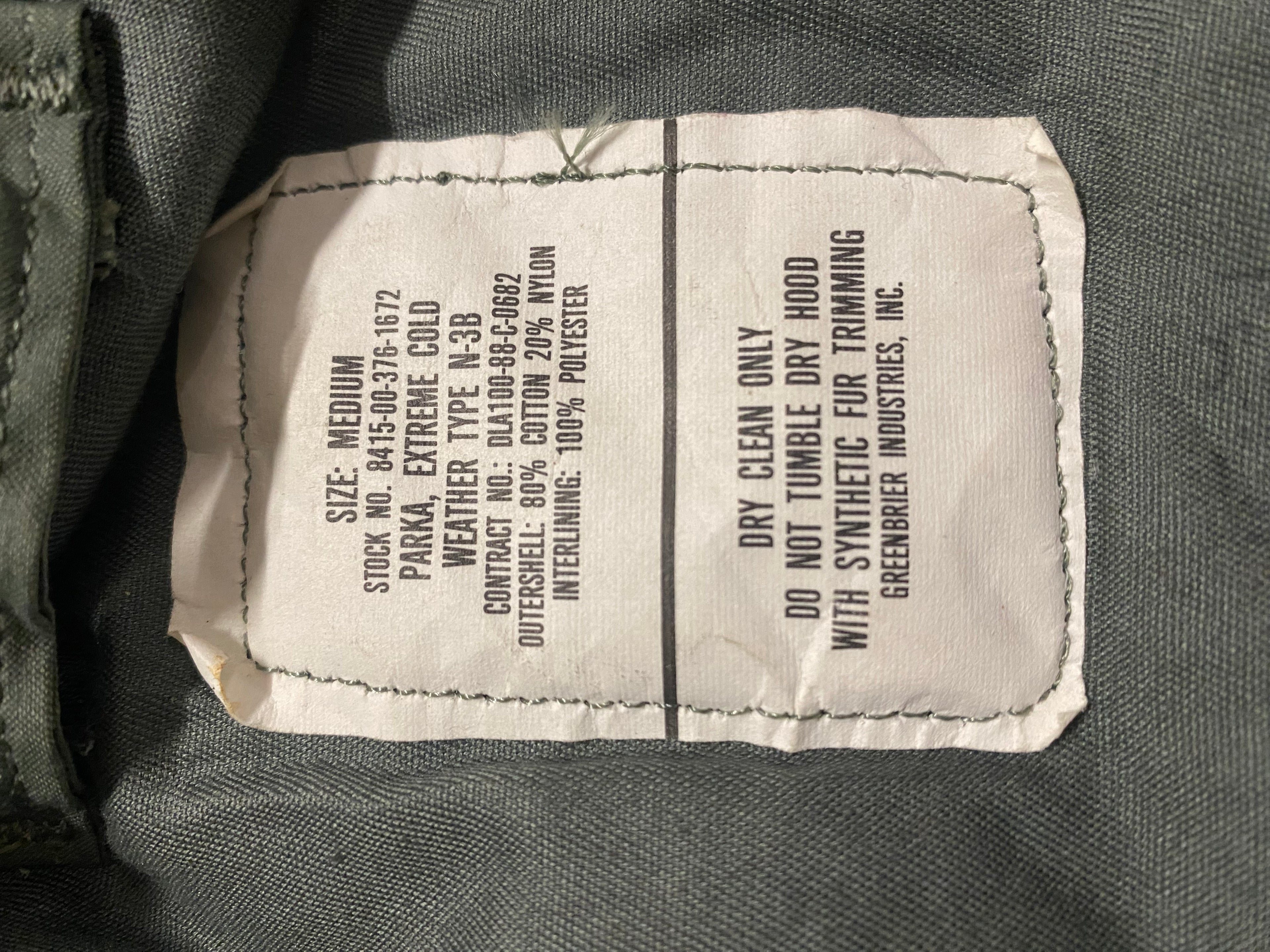 Parka N3B US Army grand froid extrême authentique: étiquette textile blanche cousue sur tissu olive gris, détails de composition et instructions d’entretien visibles.