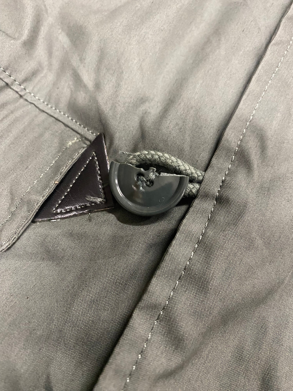 Bouton de régulation noir sur cordon épais, passant sous une patte triangulaire sur tissu gris—aération d’un Parka N3B fictif.