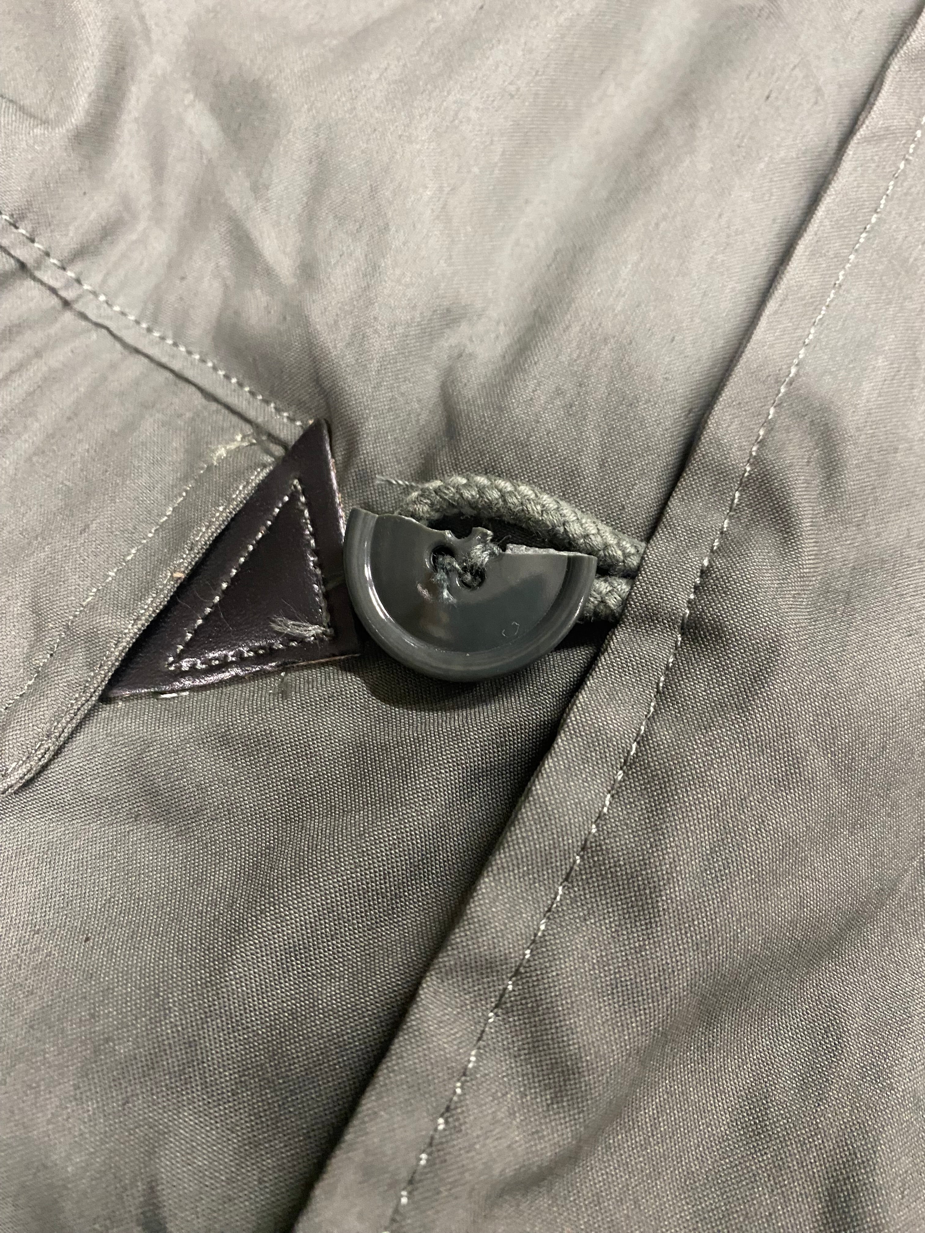 Bouton de régulation noir sur cordon épais, passant sous une patte triangulaire sur tissu gris—aération d’un Parka N3B fictif.