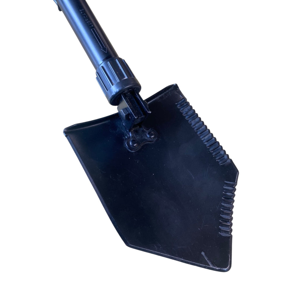 Pelle militaire pliante noire avec poignée et dentelure sur le côté, crochet de fixation visible sur un embout semi-hexagonal.