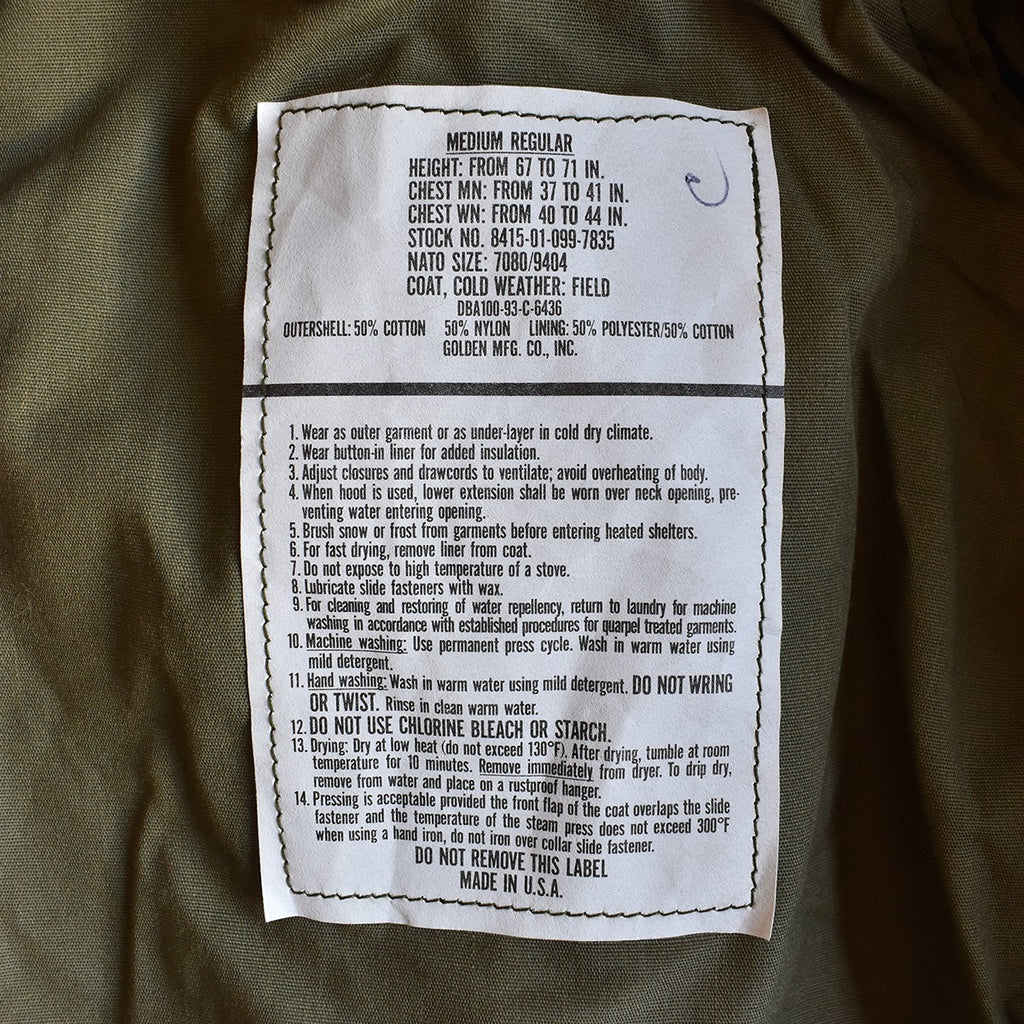 Étiquette blanche cousue sur tissu kaki illustrant les dimensions et instructions d’un manteau M-65 US Army authentique (parka militaire).