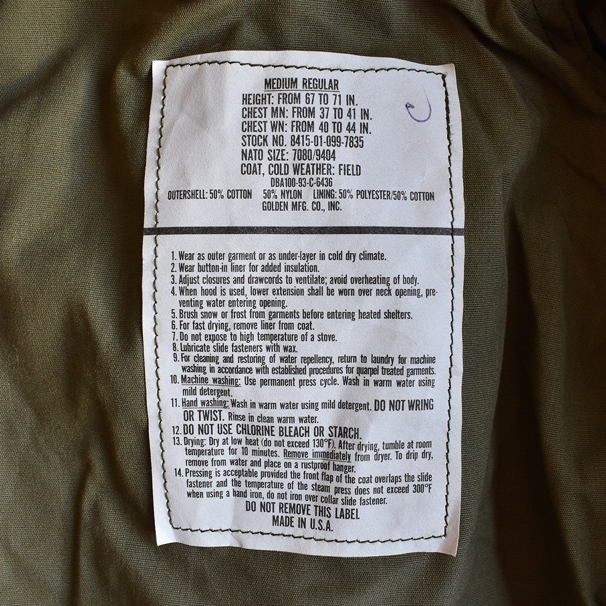 Étiquette blanche cousue sur tissu kaki illustrant les dimensions et instructions d’un manteau M-65 US Army authentique (parka militaire).